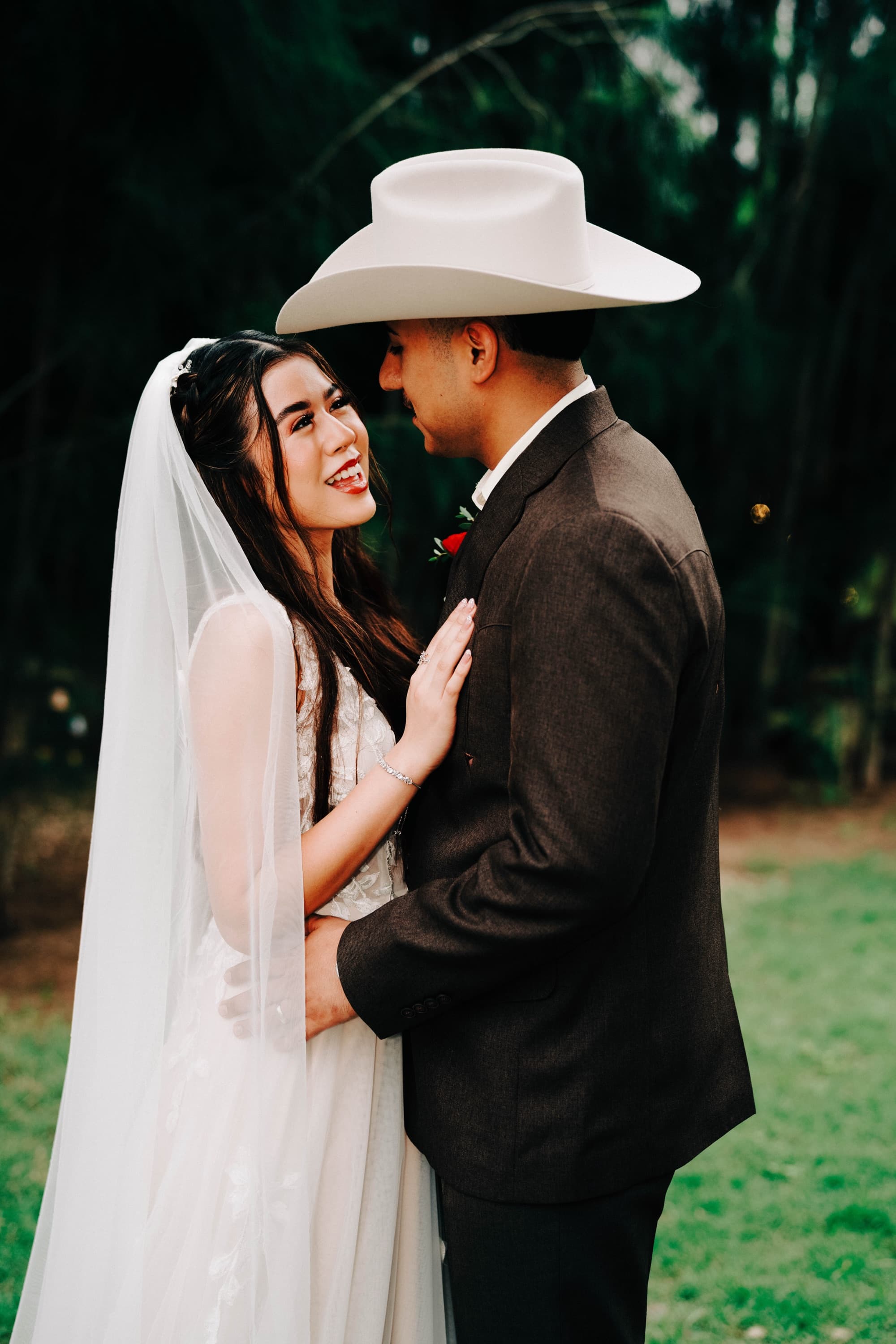 A bride laughs up at her groom, who wears a white cowboy hat, against a wall of dark green trees